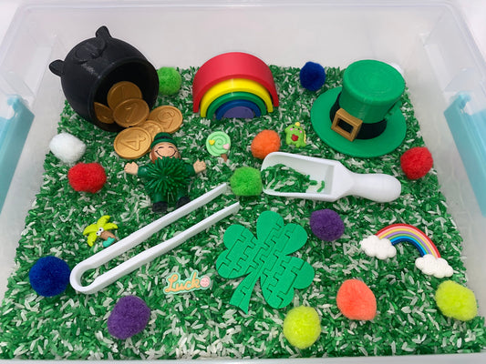 May include: A green sensory bin filled with white rice and green rice, with a variety of colorful pom-poms, a rainbow, a pot of gold coins, a leprechaun figurine, a shamrock, a green hat, and a white scoop.