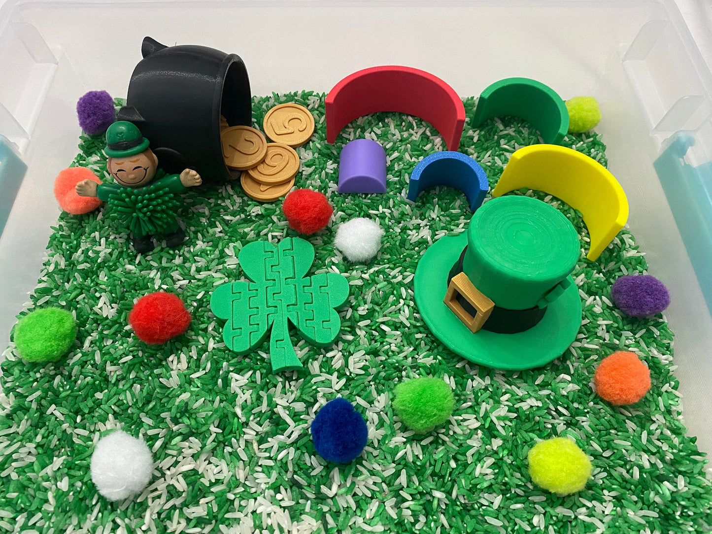 May include: A green leprechaun figurine with a pot of gold coins, a green shamrock puzzle piece, a green hat with a gold buckle, and colorful pom-poms in a sensory bin filled with green and white rice.