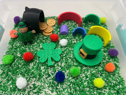 May include: A green leprechaun figurine with a pot of gold coins, a green shamrock puzzle piece, a green hat with a gold buckle, and colorful pom-poms in a sensory bin filled with green and white rice.