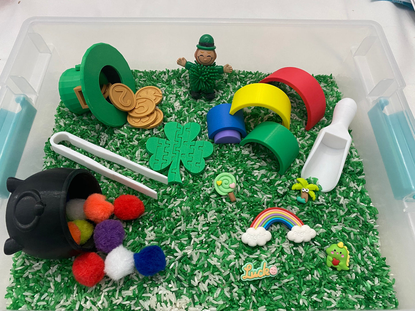 May include: A green sensory bin filled with green and white rice. The bin contains a variety of toys including a leprechaun, a pot of gold, a shamrock, a rainbow, and a scoop.