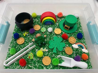 May include: A clear plastic container filled with green rice, green pom-poms, and other St. Patrick's Day themed items, including a leprechaun figurine, a pot of gold, a rainbow, a shamrock, and a green hat. The numbers 1, 4, 6, and 7 are also included.