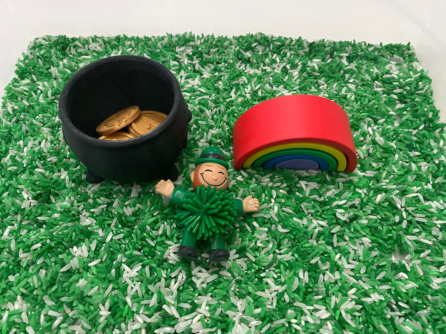 May include: A black cauldron filled with gold coins, a green leprechaun toy, and a rainbow toy on a green and white rice sensory bin.