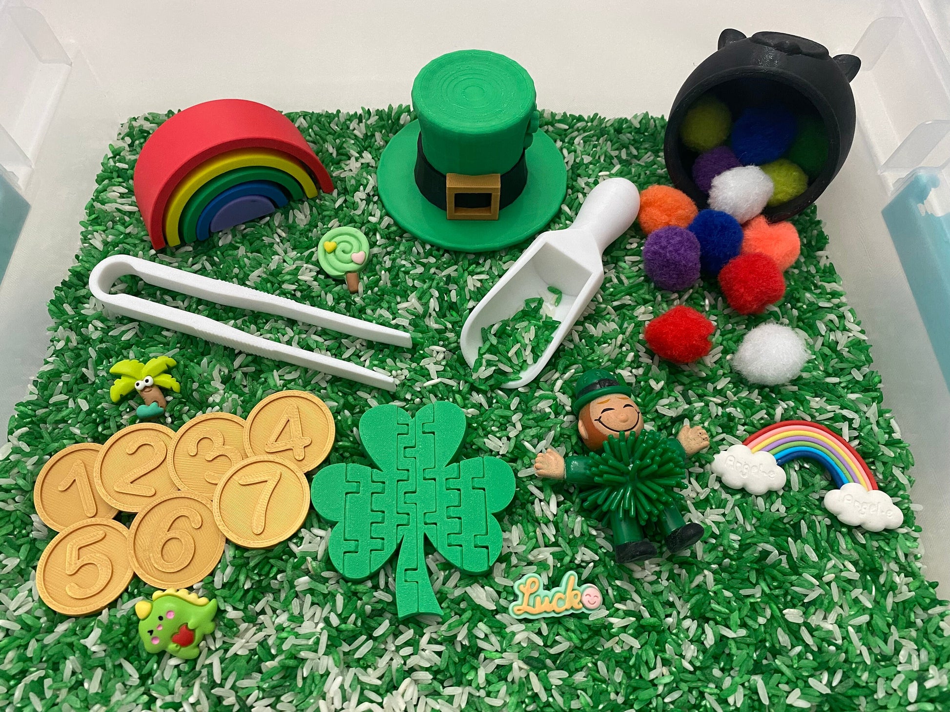 May include: A green and white sensory bin filled with rice, featuring a green leprechaun figurine, a green shamrock puzzle, a rainbow, a pot of gold, and a green hat.