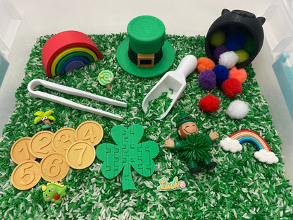 May include: A green and white sensory bin filled with rice, featuring a green leprechaun figurine, a green shamrock puzzle, a rainbow, a pot of gold, and a green hat.