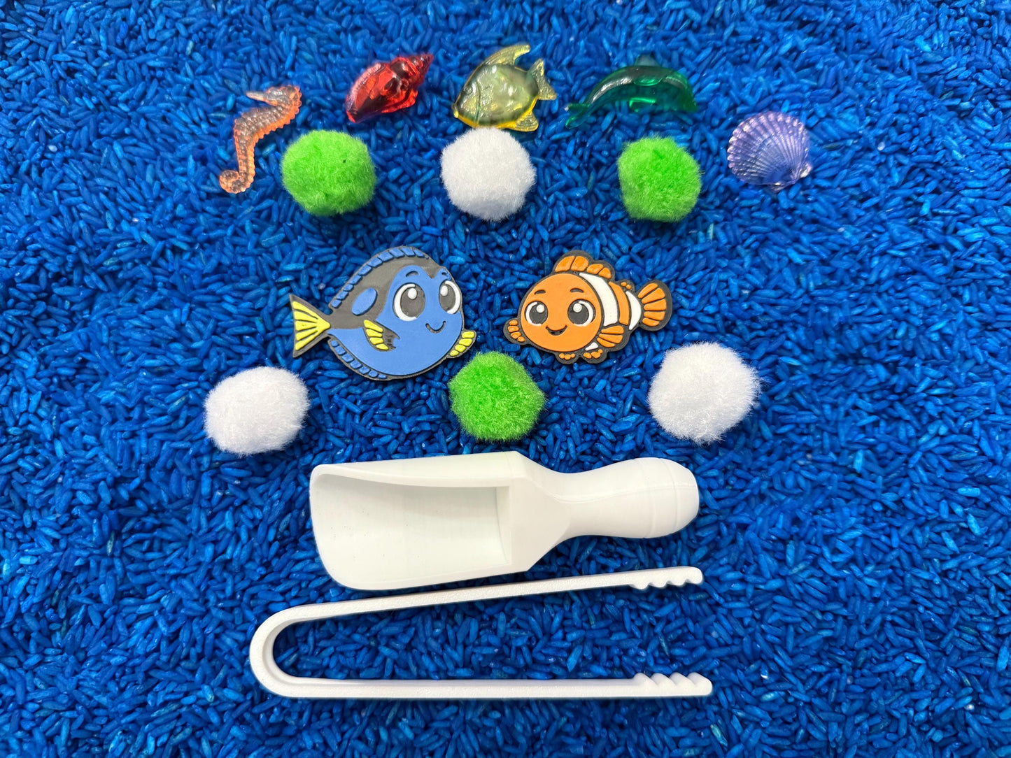 An ocean-themed sensory bin with blue rice, featuring toy fish, a seahorse, a seashell, green pom-poms, and white scoops and tongs. The toys are arranged on blue rice, creating a playful and engaging scene.