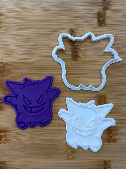 May include: Two cookie cutters, one purple and one white, shaped like the Pokemon Ghost type, Gengar. The cutters are on a wooden surface.