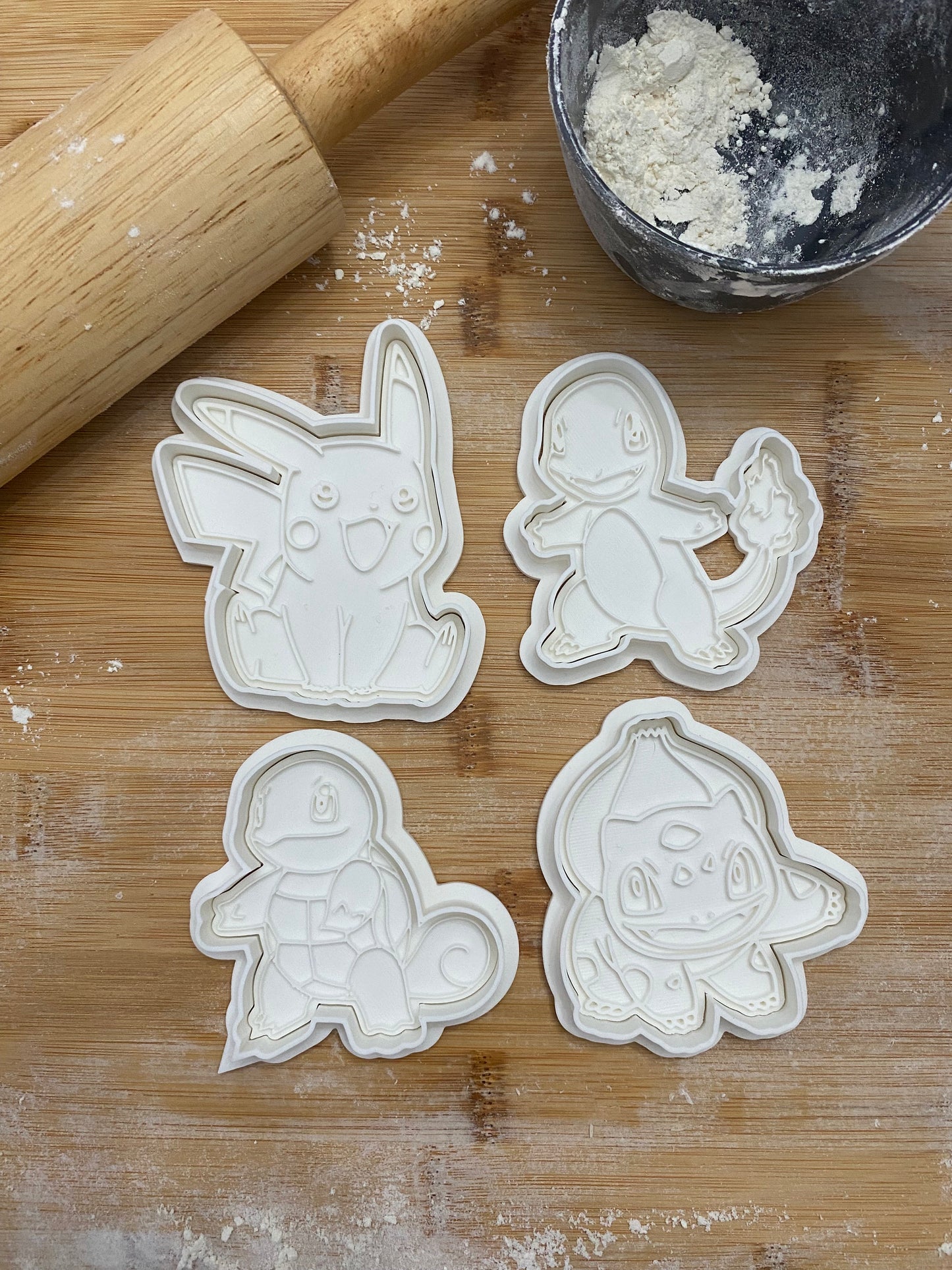 May include: Four white plastic cookie cutters shaped like Pokemon characters: Pikachu, Charmander, Squirtle, and Bulbasaur. The cutters are on a wooden surface with white flour scattered around them.