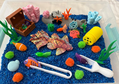 May include: A blue sensory bin filled with blue rice, featuring a variety of sea-themed toys, including a treasure chest, fish, coral, and seaweed. There are also colorful pom-poms and a pair of tweezers.