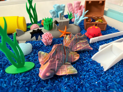 May include: A blue sensory bin filled with blue rice, featuring a variety of colorful plastic toys, including a pink fish, a gray shark, a red crab, a yellow barrel, a blue submarine, and green seaweed.