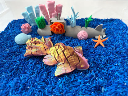 May include: A blue and white submarine toy with orange details, surrounded by coral, seaweed, and starfish toys, all sitting on a bed of blue rice.