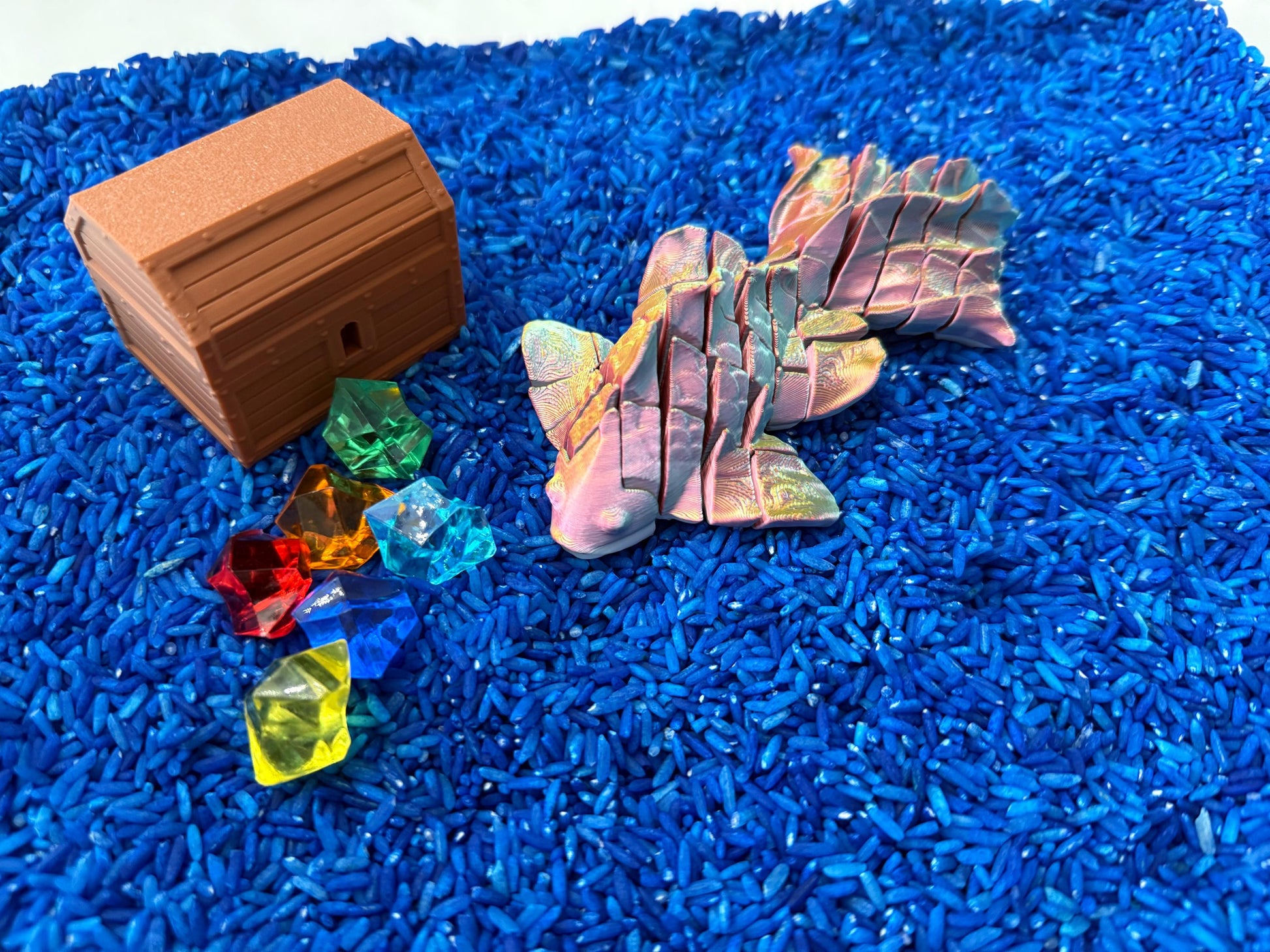 May include: A wooden treasure chest with a lid open, revealing a pile of colorful gems. The chest is sitting on a bed of blue rice, along with a iridescent origami fish.