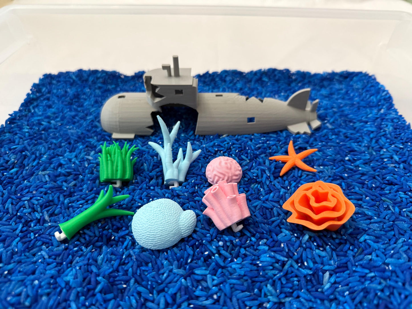May include: A gray toy submarine broken into two pieces, surrounded by blue rice. There are also several other toy sea creatures, including a starfish, coral, seaweed, and a sea shell.