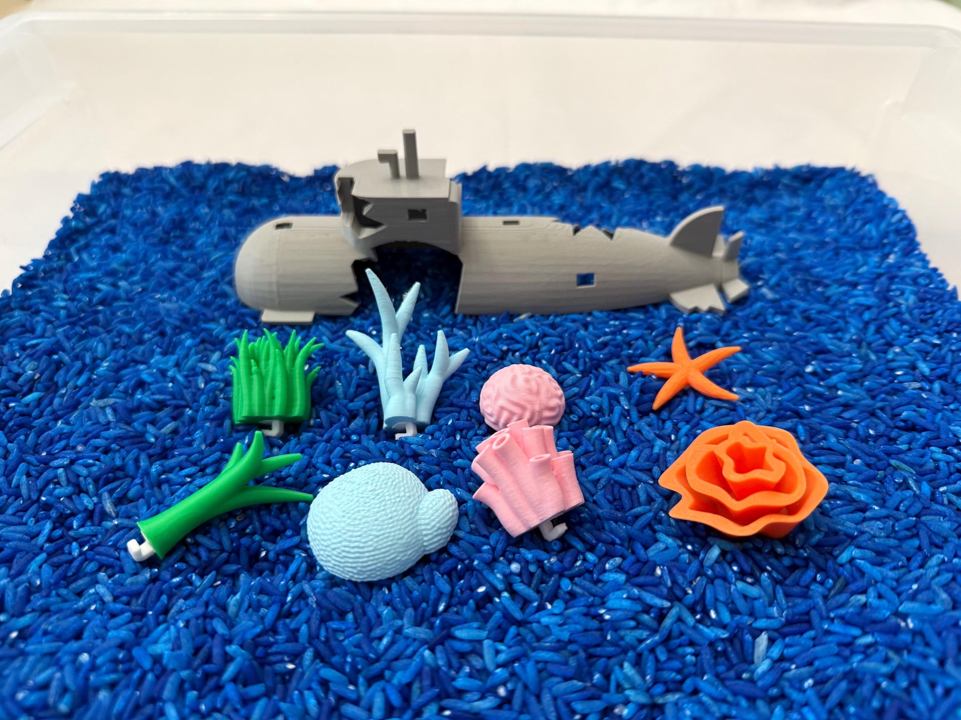 May include: A gray toy submarine broken into two pieces, surrounded by blue rice. There are also several other toy sea creatures, including a starfish, coral, seaweed, and a sea shell.