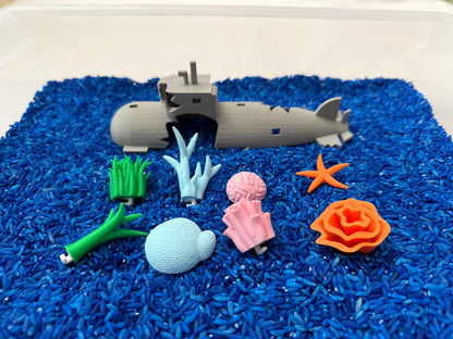 May include: A gray toy submarine broken into two pieces, surrounded by blue rice. There are also several other toy sea creatures, including a starfish, coral, seaweed, and a sea shell.