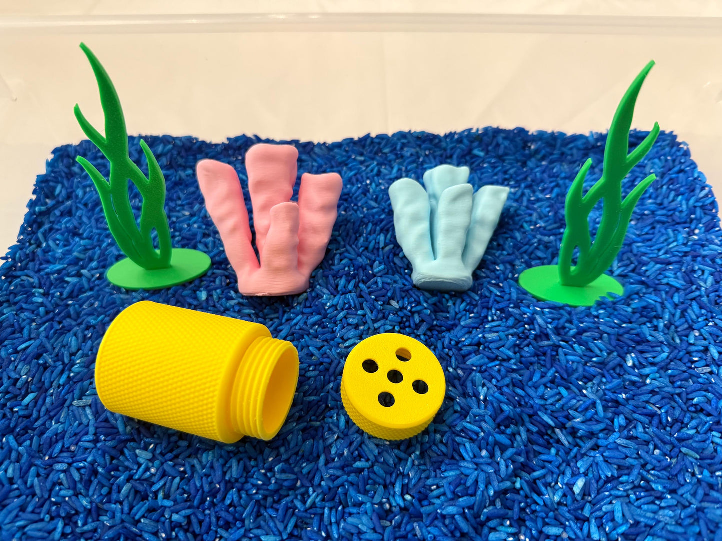 May include: A blue sensory bin filled with blue rice. There are two green plastic seagrass plants, two coral pieces, one yellow plastic container, and one yellow plastic lid with holes in it.