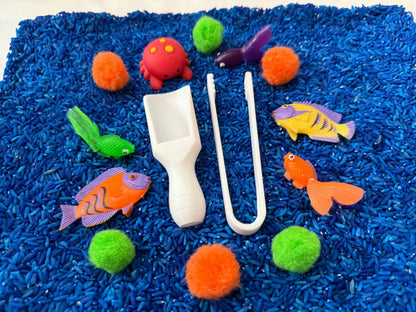 May include: A blue sensory bin filled with blue rice, featuring a variety of colorful sea creatures, including a red crab, a yellow fish, an orange fish, a green fish, and a purple fish. The bin also includes two white plastic tools, a scoop and a pair of tongs, and several colorful pom-poms.