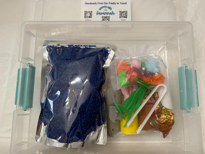 May include: A clear plastic storage container filled with blue sand, a bag of colorful plastic flowers, and a pair of tweezers. The container is labeled "Handmade From Our Family to Yours" with a logo that says "Sweetly Made by Savannah".