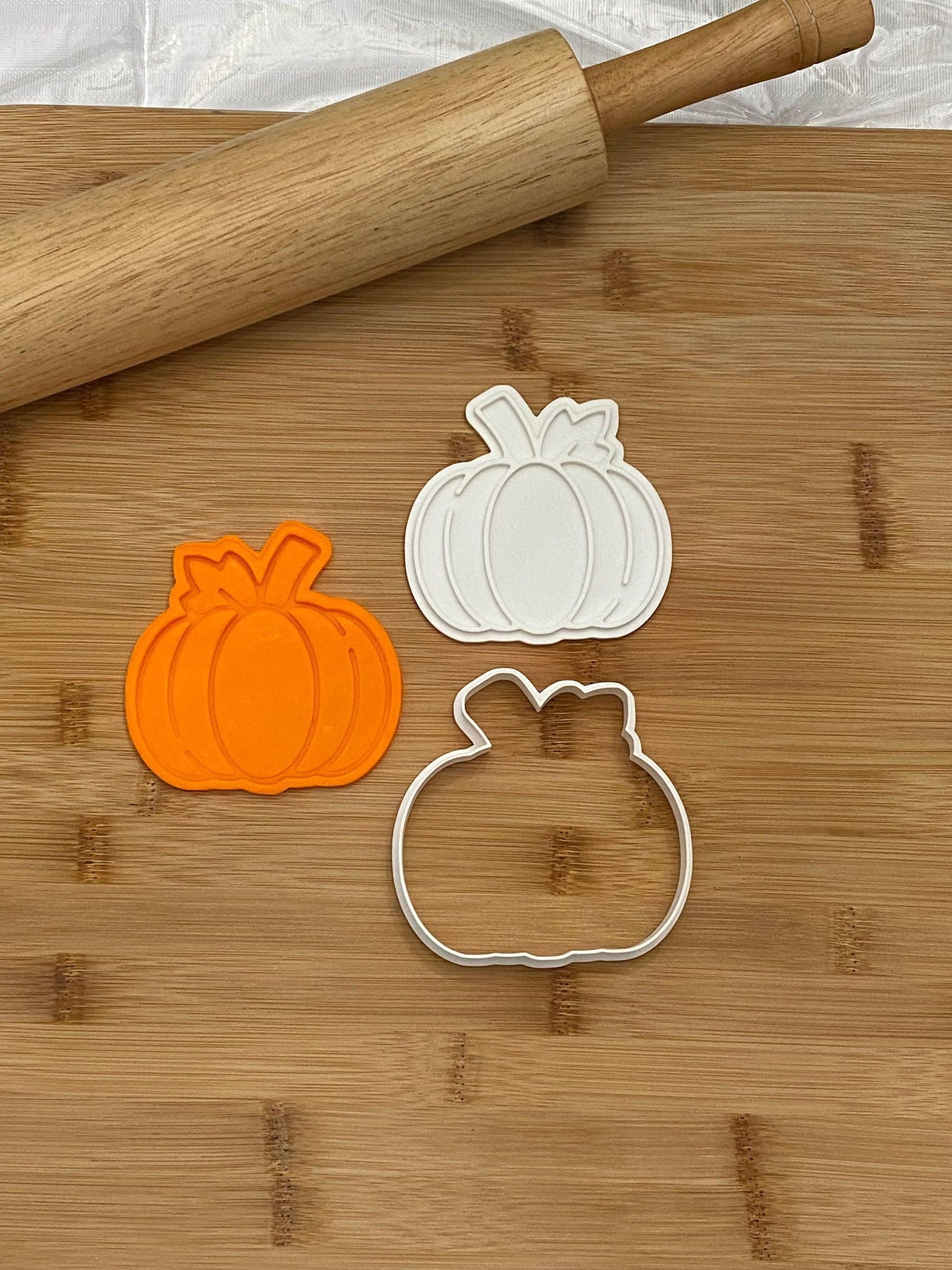 May include: Two white plastic pumpkin cookie cutters and one orange plastic pumpkin cookie cutter on a wooden cutting board with a wooden rolling pin.