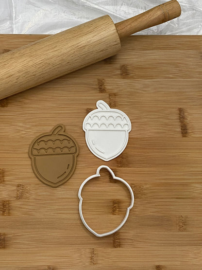 May include: Two white plastic acorn-shaped cookie cutters on a wooden cutting board with a wooden rolling pin.