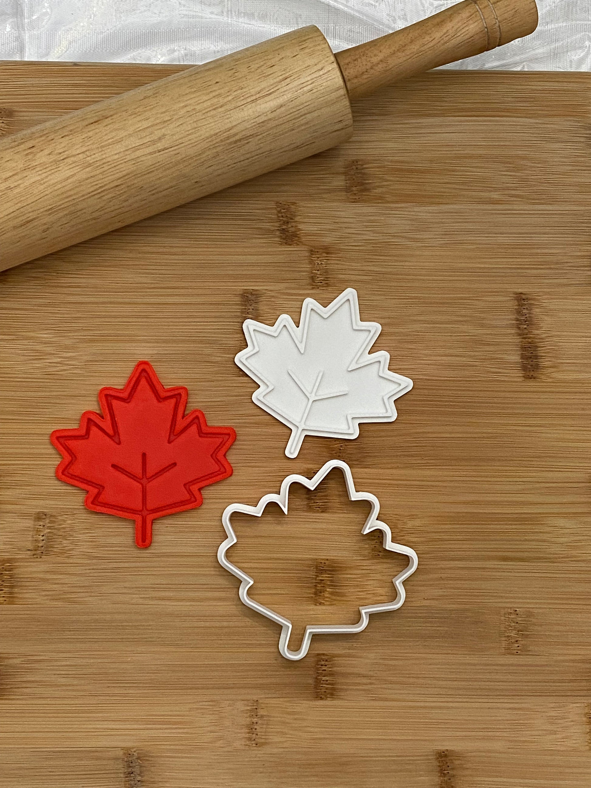 May include: Three maple leaf cookie cutters, two white and one red, on a wooden cutting board with a rolling pin.