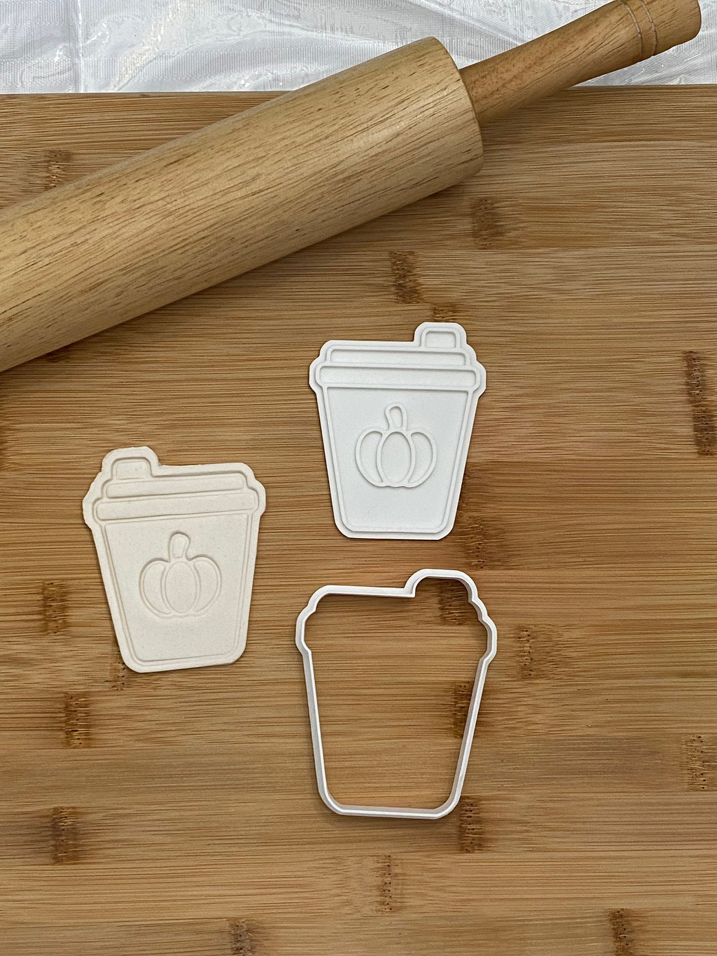 May include: Three white plastic cookie cutters shaped like coffee cups with a pumpkin design. The cutters are on a wooden cutting board with a rolling pin.