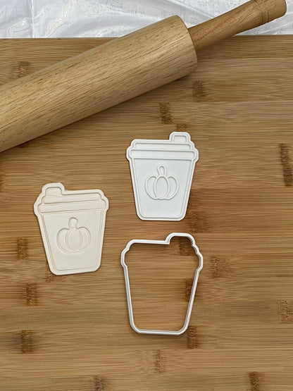 May include: Three white plastic cookie cutters shaped like coffee cups with a pumpkin design. The cutters are on a wooden cutting board with a rolling pin.